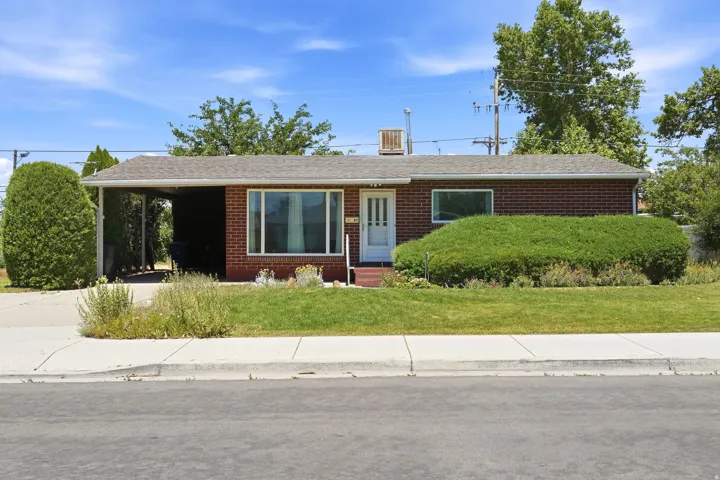 Brick exterior property featuring a covered carport, established lawn, mature landscaping, and a white entry door