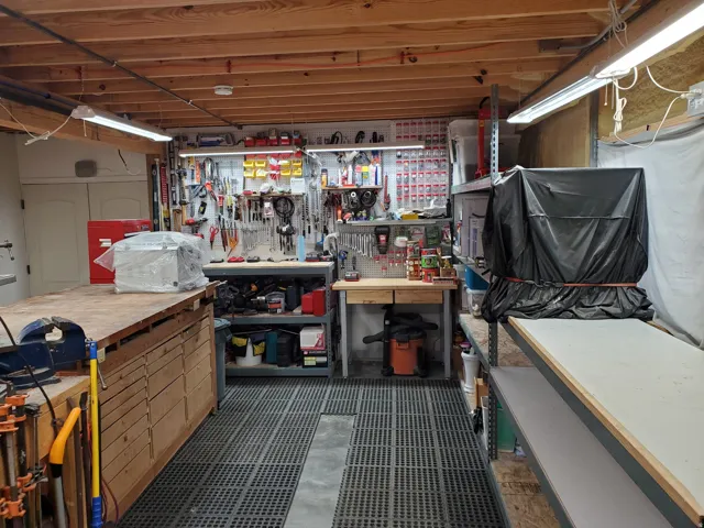 Workshop space featuring exposed wood ceiling joists, overhead fluorescent lighting, a built-in workbench with storage, and a work surface with open shelving