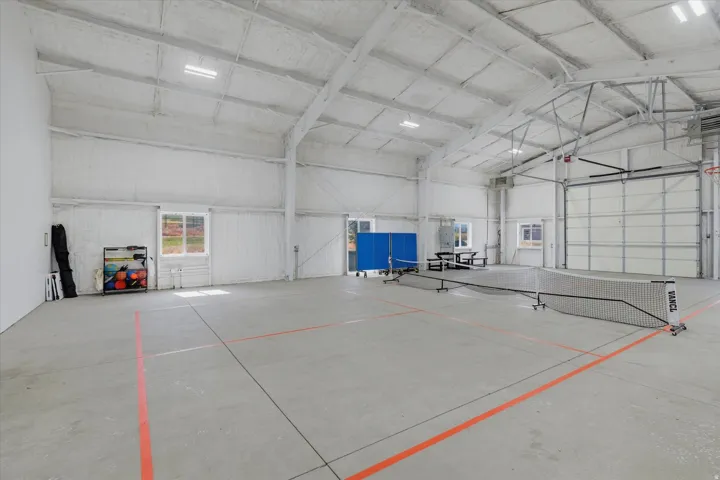 Spacious indoor court featuring high ceilings with exposed trusses, concrete flooring with painted lines, multiple windows, and a large roll-up door