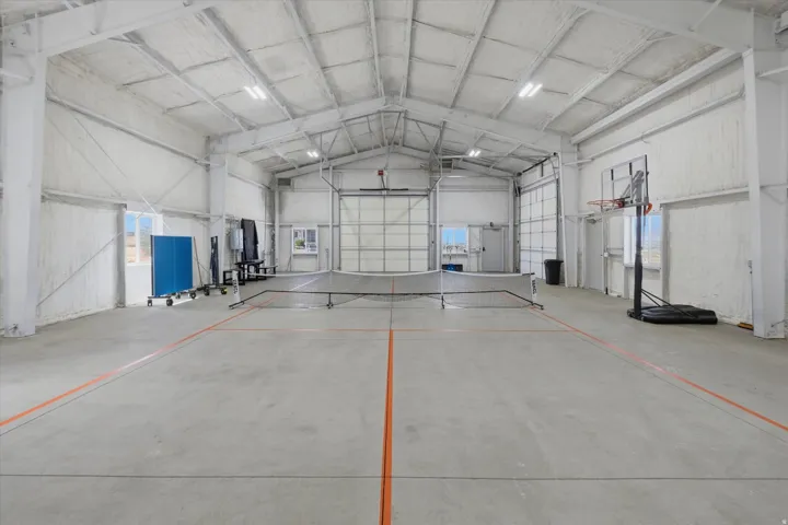 Spacious indoor recreational facility featuring concrete flooring with court line markings, high ceilings with exposed trusses, multiple overhead lighting fixtures, and large rollup doors