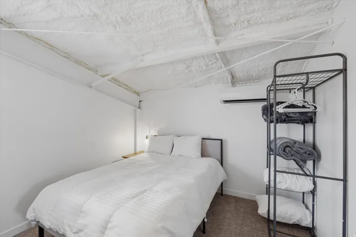 Bedroom featuring a wall-mounted AC unit, exposed beam ceiling with spray foam insulation, and a built-in wall sconce