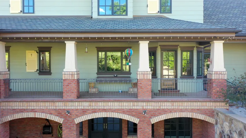 Multi-level exterior featuring a covered porch with stone columns and wrought iron railing, brick archways, multiple windows with dark trim, and light green siding