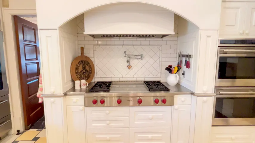 Kitchen featuring a stainless steel range with red knobs, integrated double ovens, white cabinetry, subway tile backsplash, and a wall-mounted pot filler