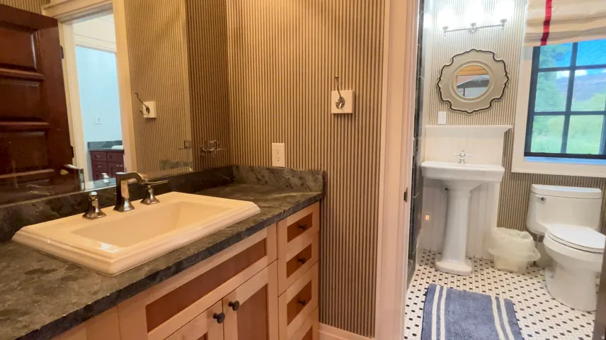 Two distinct vanity areas featuring a light wood-finish vanity with a dark stone countertop and an undermount sink, alongside a classic pedestal sink