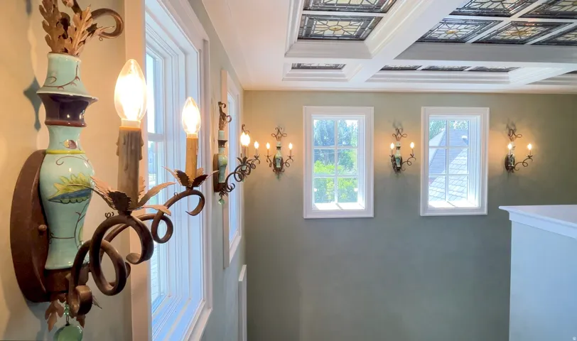 Ornate sconces illuminate a hallway featuring multiple windows, a coffered ceiling with decorative panels, and a painted wall finish
