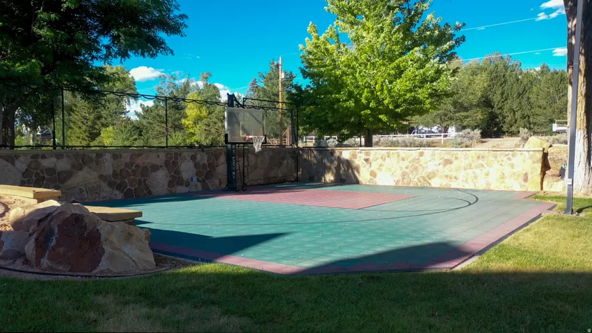 Outdoor sport court featuring a basketball hoop, multi-colored modular tile surface, and a stone retaining wall