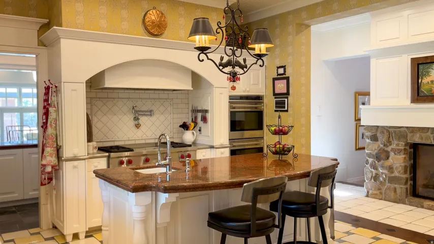 Gourmet kitchen featuring a large central island with a polished countertop and seating, professional-grade range with pot filler, custom range hood, double wall ovens, and a stone fireplace surround