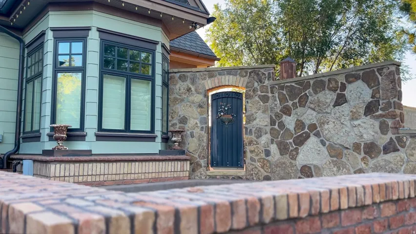 Stone wall with arched entryway featuring an ornate dark-colored door, brick coping, and a bay window with black trim