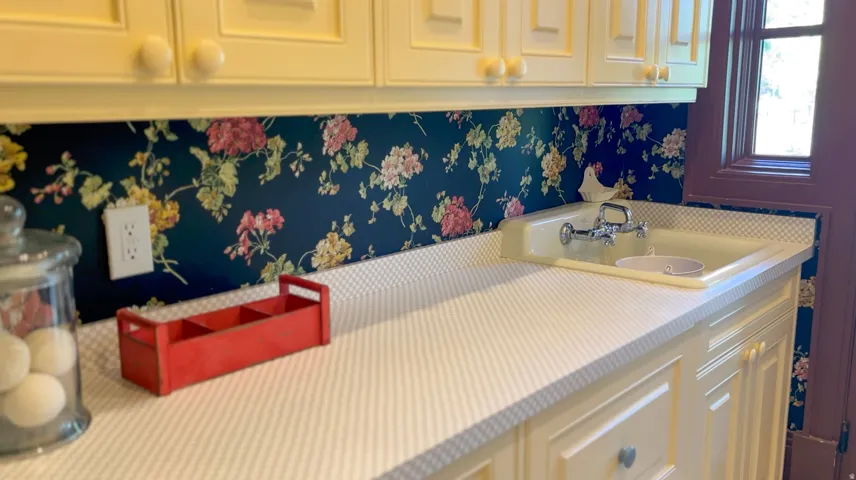 Cream cabinetry with round knobs, floral wallpaper backsplash, light-toned countertop, and an integrated sink with a chrome faucet