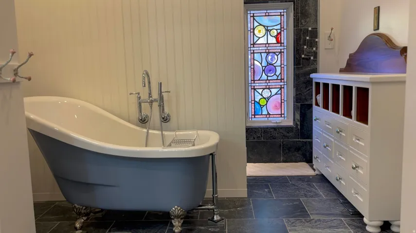 Freestanding clawfoot tub with polished chrome fixtures, slate tile flooring, and stained glass window feature