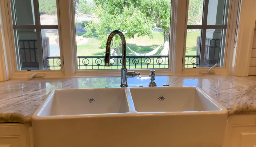 Double basin farmhouse sink with a polished chrome gooseneck faucet, set within a light-toned stone countertop, framed by two windows featuring decorative ironwork