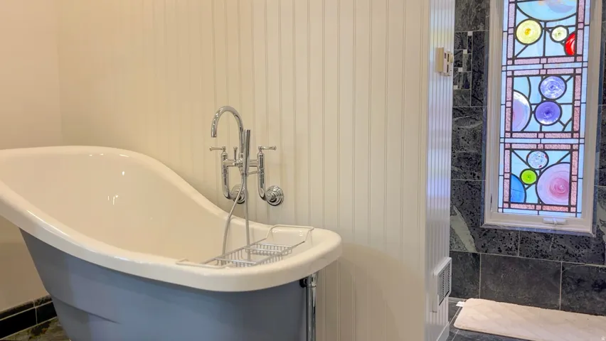 Freestanding bathtub with chrome fixtures, beadboard wall paneling, and a stained glass window