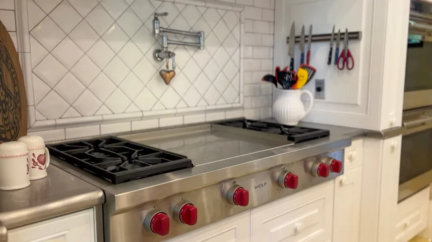 Professional-grade Wolf range with red control knobs, integrated griddle, stainless steel countertop, white cabinetry with recessed panel doors, and a white subway tile backsplash with a diagonal pattern