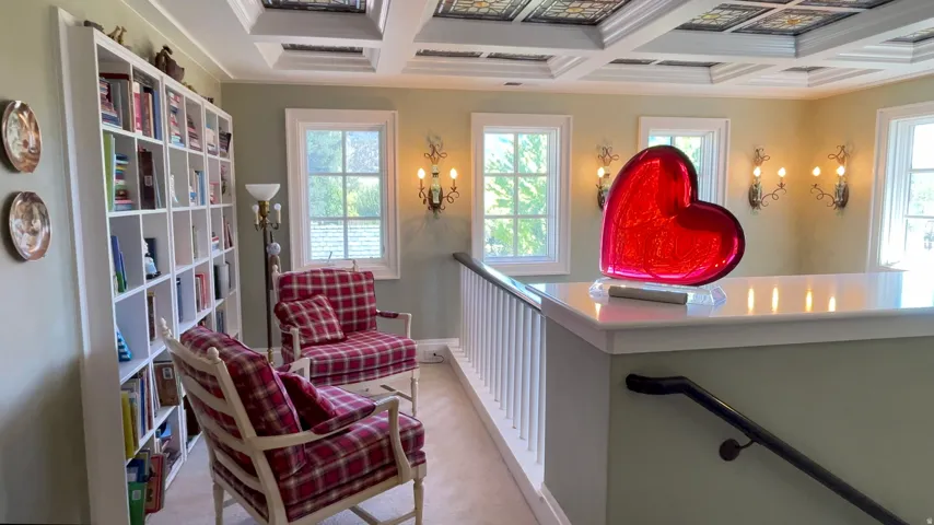 Upper-level landing featuring a coffered ceiling with decorative panels, built-in shelving, white trim windows, wall sconces, and a white railing with a dark-toned handrail