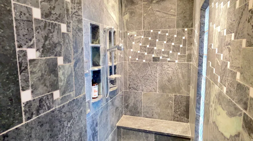 Shower enclosure featuring large format stone-finish tile, decorative tile accents, built-in shower bench, recessed storage niches, and a handheld shower fixture