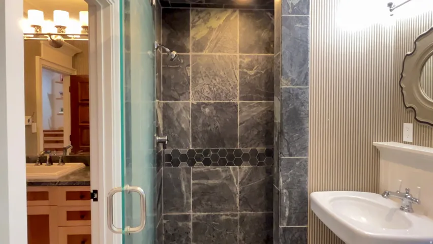 Shower enclosure featuring large format stone-finish tile with a hexagonal mosaic accent band, a glass door with a polished chrome handle, and a wall-mounted showerhead