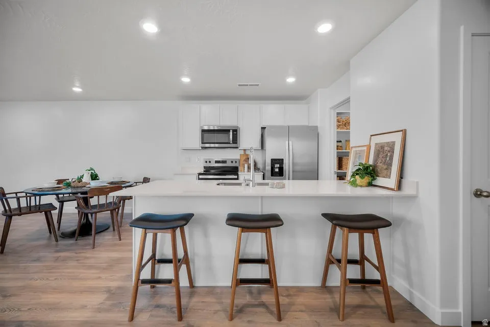 Modern kitchen featuring white cabinetry, stainless steel appliances, a breakfast bar with seating, wood-finish flooring, and recessed lighting