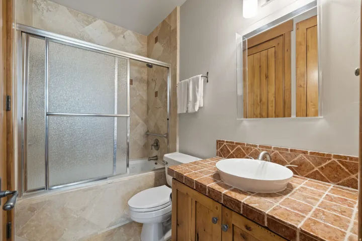 Bathroom featuring a tile-surround tub/shower with sliding glass doors, a white toilet, and a wood-finish vanity with a tiled countertop and vessel sink