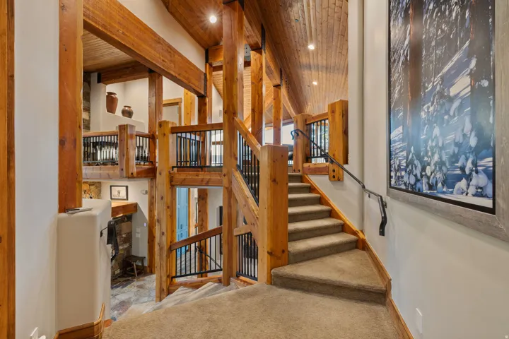 Multi-level interior featuring exposed timber beams, wood-finish vaulted ceilings, carpeted staircase with dark metal handrail, and a stone-clad fireplace