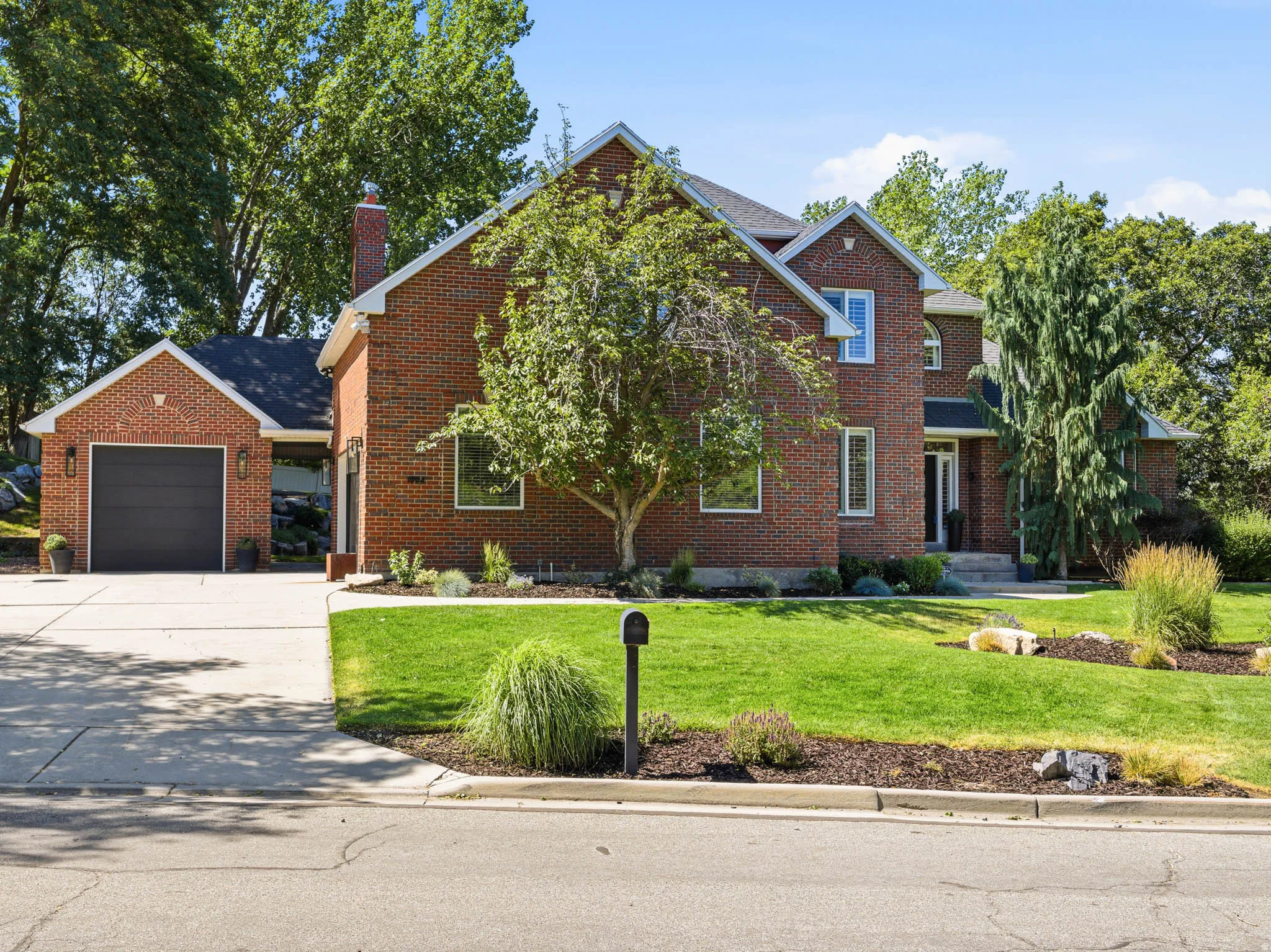 Brick exterior residence featuring a two-car garage with a dark gray door, a neatly manicured lawn, established trees, and a concrete driveway
