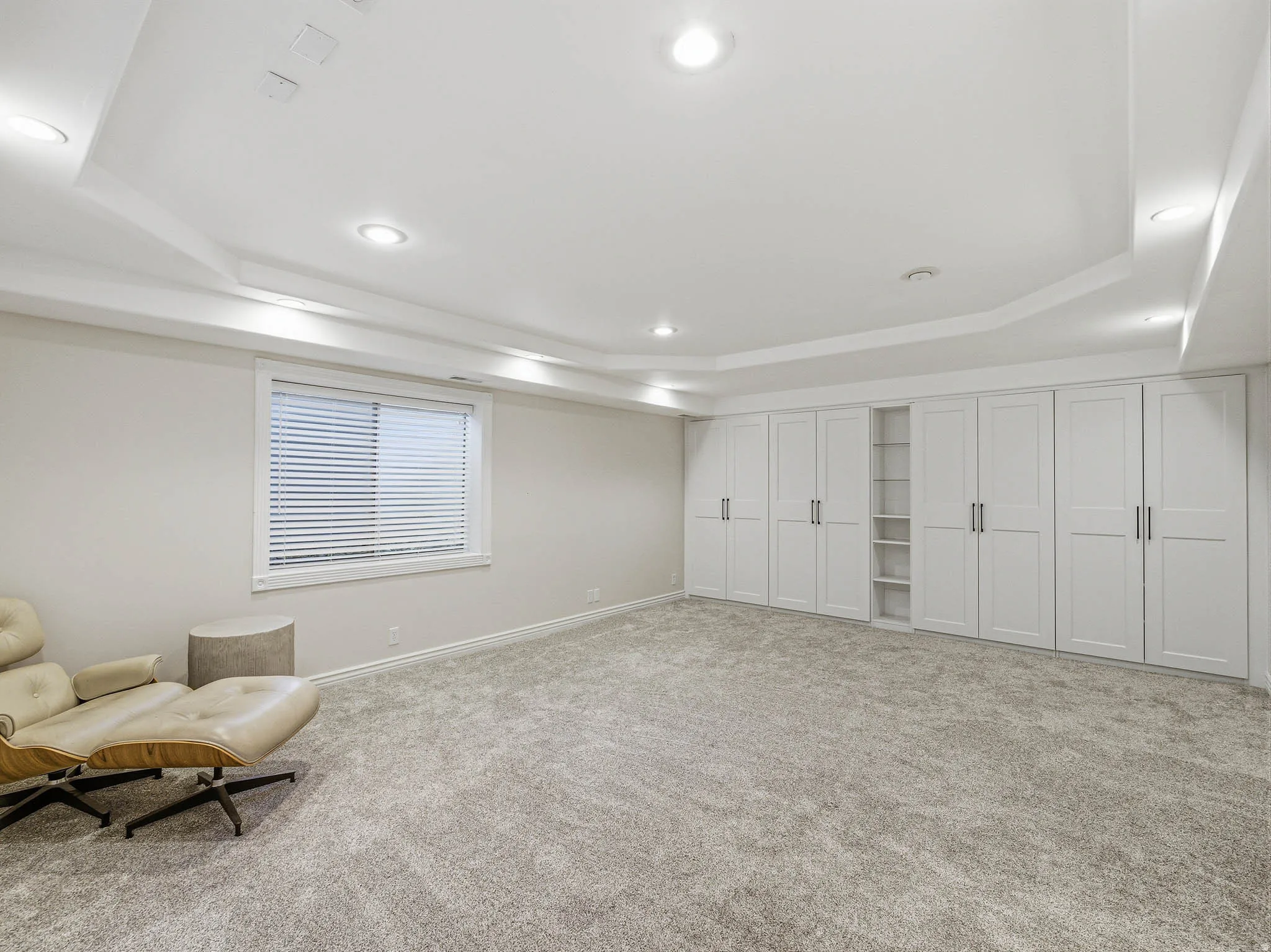 Spacious room featuring extensive built-in cabinetry with brushed nickel hardware, soft gray carpet, tray ceiling with recessed lighting, and a single window with horizontal blinds