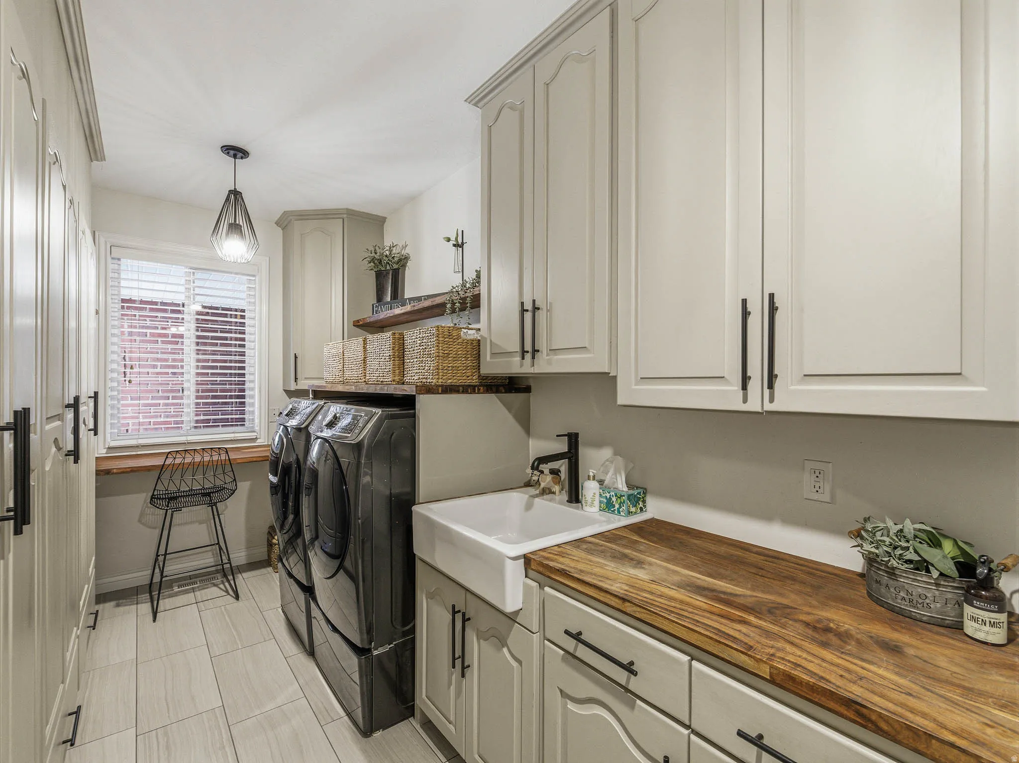Laundry room featuring extensive light-toned cabinetry, light-finish tile flooring, a farmhouse sink with a black faucet, and a wood-finish countertop