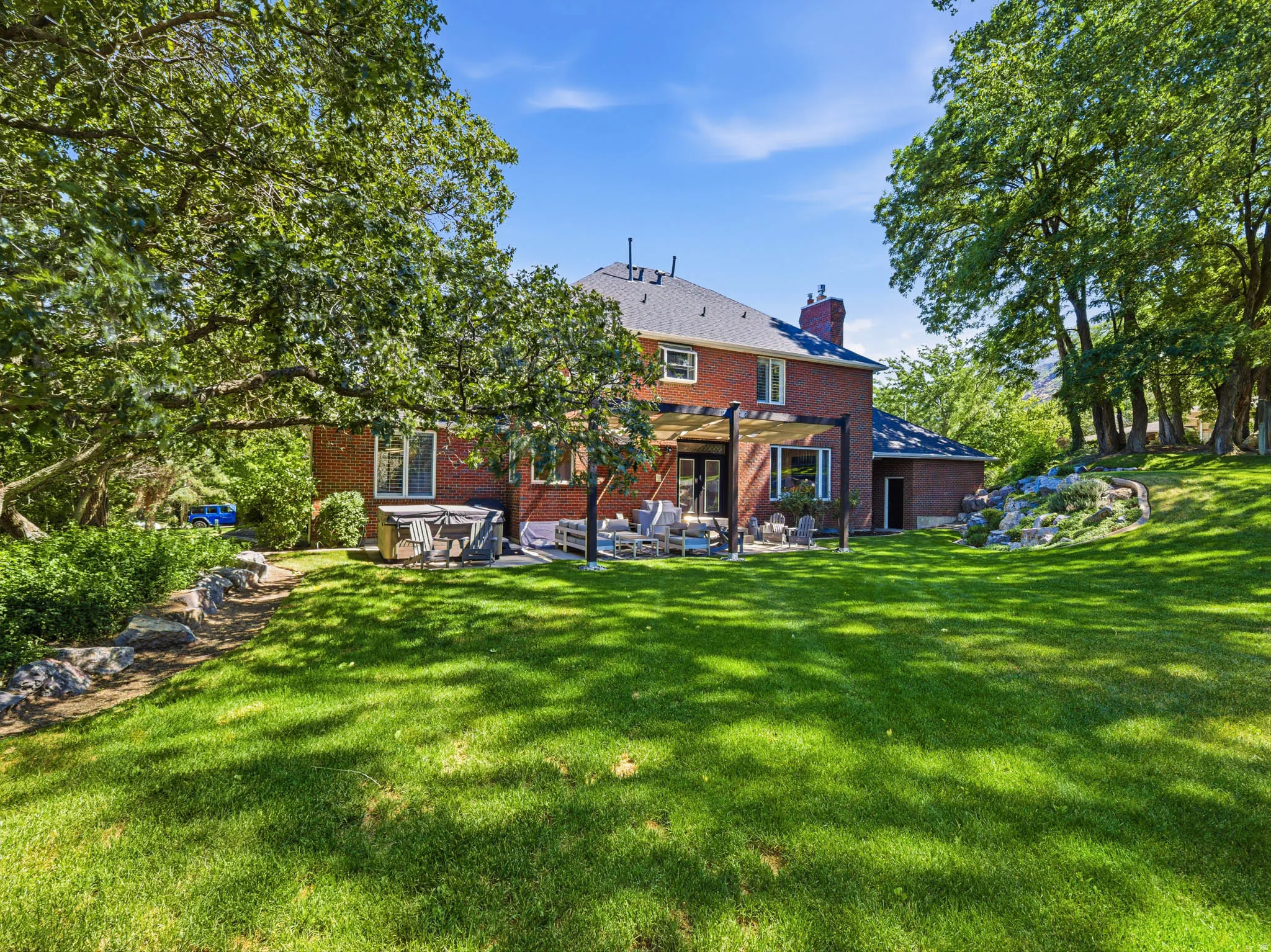 Spacious backyard with mature trees, a red brick exterior, a stone patio with a pergola, and a terraced rock garden
