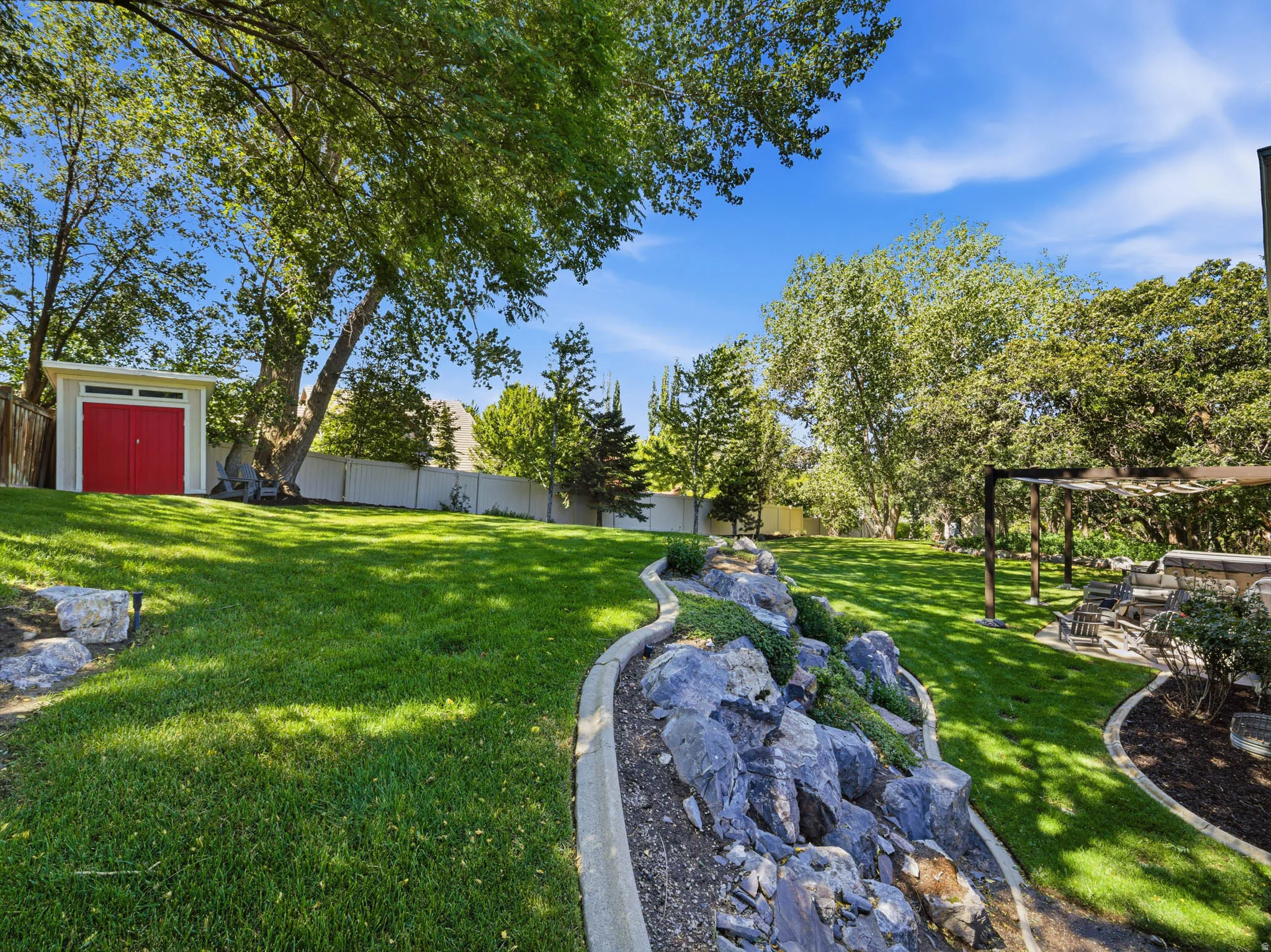 Spacious backyard featuring a manicured lawn, established trees, a white picket fence, a modern storage shed with red double doors, and a decorative rock garden