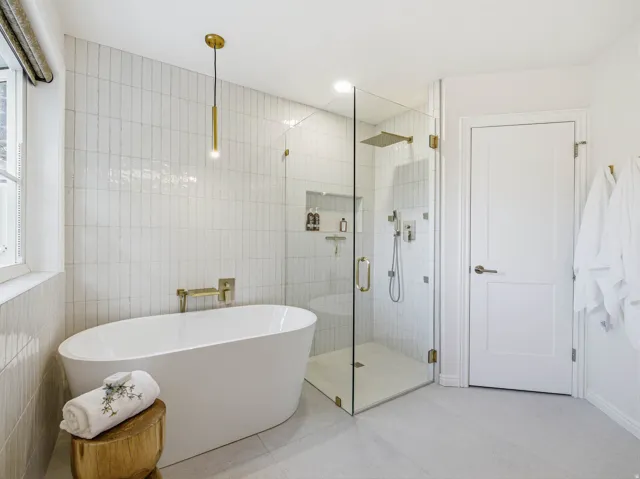 Freestanding soaking tub with a wall-mounted brass faucet, glass-enclosed shower with a rainfall showerhead, and vertical subway tile accents