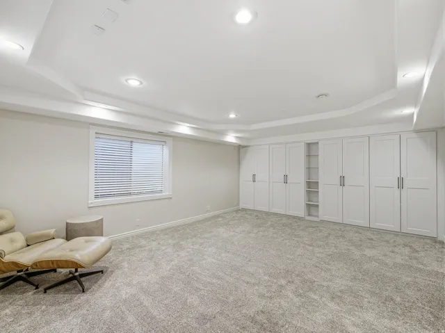 Spacious room featuring extensive built-in cabinetry with brushed nickel hardware, soft gray carpet, tray ceiling with recessed lighting, and a single window with horizontal blinds