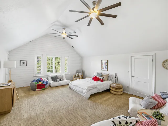 Vaulted ceiling room featuring two contemporary ceiling fans, white shiplap accent wall, multiple windows with plantation shutters, and light brown carpeting