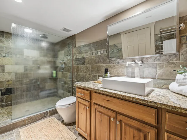 Bathroom featuring natural stone tile walls and flooring, a wood-finish vanity with granite countertop, a rectangular vessel sink with wall-mounted faucet, and a glass-enclosed shower