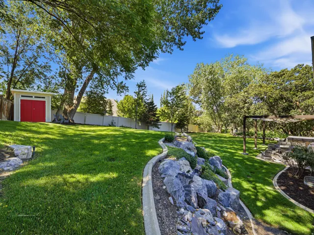 Spacious backyard featuring a manicured lawn, established trees, a white picket fence, a modern storage shed with red double doors, and a decorative rock garden