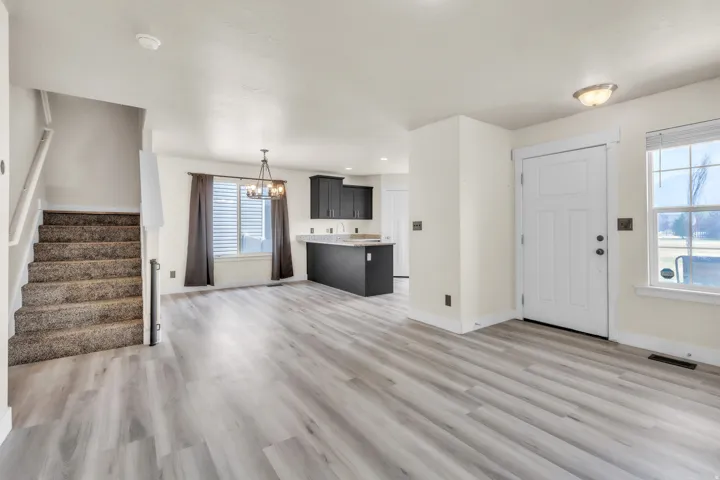 Spacious main living area featuring light wood-finish flooring, a white paneled entry door, and a staircase with textured carpet