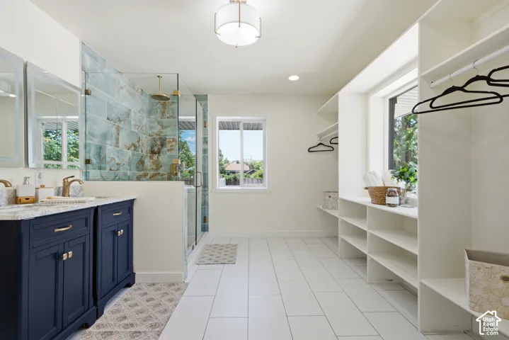 Expansive bathroom featuring a double vanity with navy cabinetry, a glass-enclosed shower with decorative tile, white tile flooring, and integrated open shelving and hanging storage