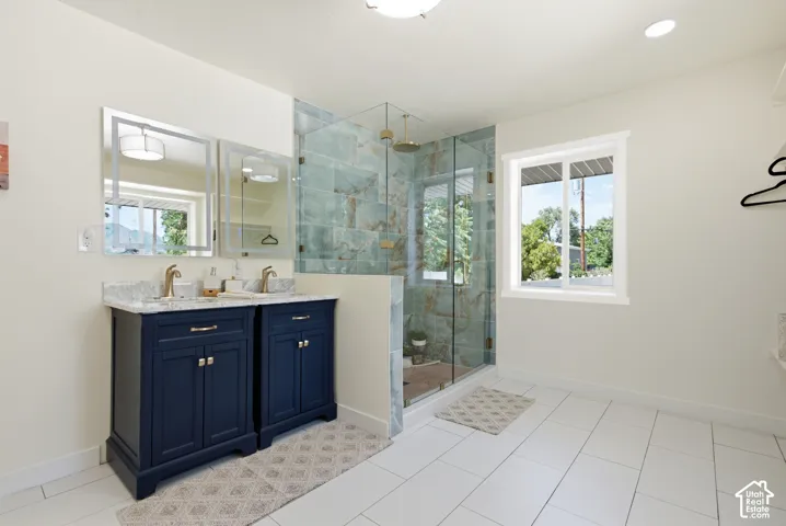 Spacious bathroom featuring a dual vanity with navy cabinetry and gold-toned fixtures, a glass-enclosed shower with decorative wall tiling, and white rectangular floor tiles