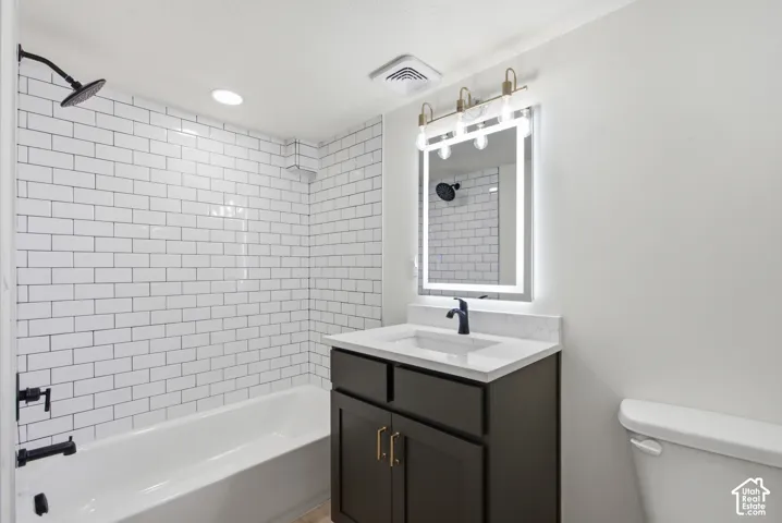 Bathroom featuring white subway tile surround, matte black shower fixtures, a dark wood-finish vanity with a white countertop and integrated sink, and an illuminated mirror with brass-finish lighting