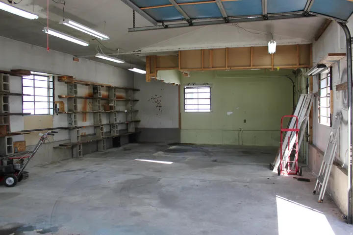 Concrete flooring, multiple fluorescent light fixtures, exposed ceiling joists, two windows with security bars, and built-in shelving units