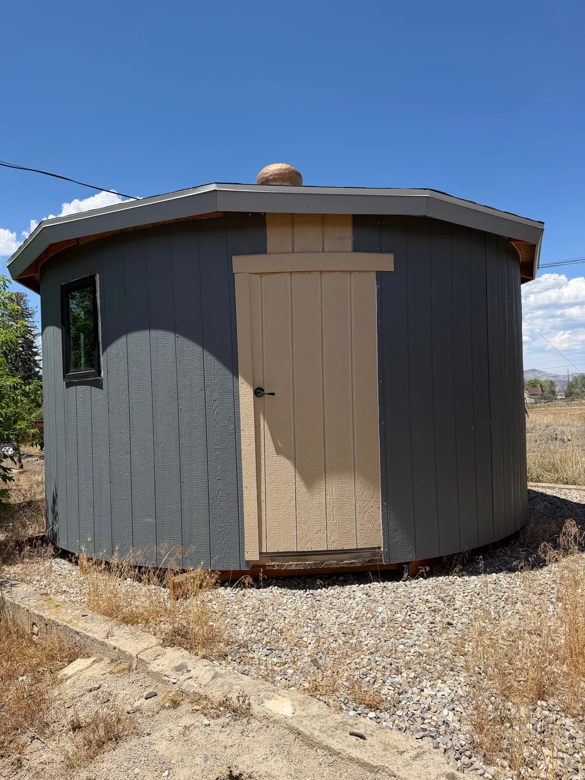 Octagonal outbuilding with dark gray siding and beige trim, featuring a single window and a solid door
