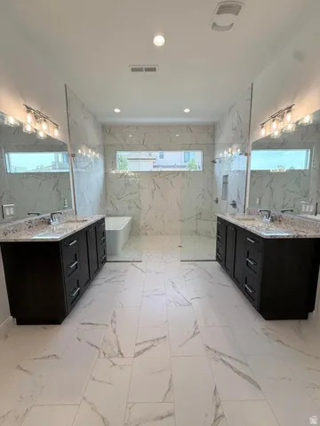 Spacious bathroom featuring marble-look tile flooring, dual dark wood-finish vanities with light stone countertops, and a large walk-in shower with a freestanding tub