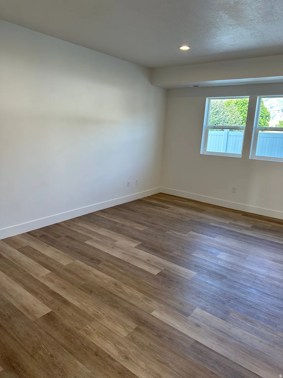 Spacious room featuring wood-finish flooring, white baseboards, recessed ceiling lighting, and two casement windows with white frames
