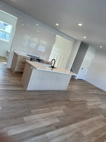 Modern kitchen featuring a central island with an integrated sink and matte black faucet, light-toned wood-finish cabinetry, white subway tile backsplash, and quartz countertops
