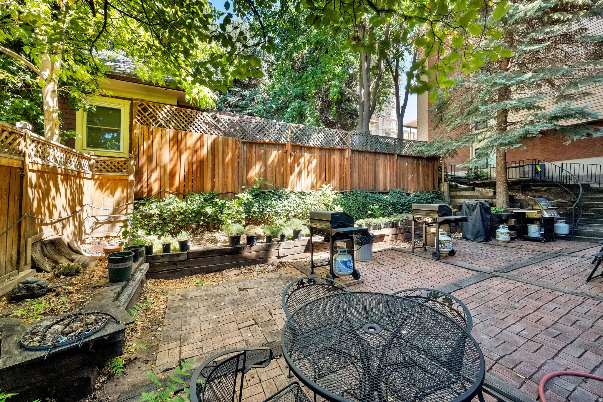 Expansive brick paver patio featuring a perimeter wood fence, tiered landscaping, mature trees, and a visible building exterior