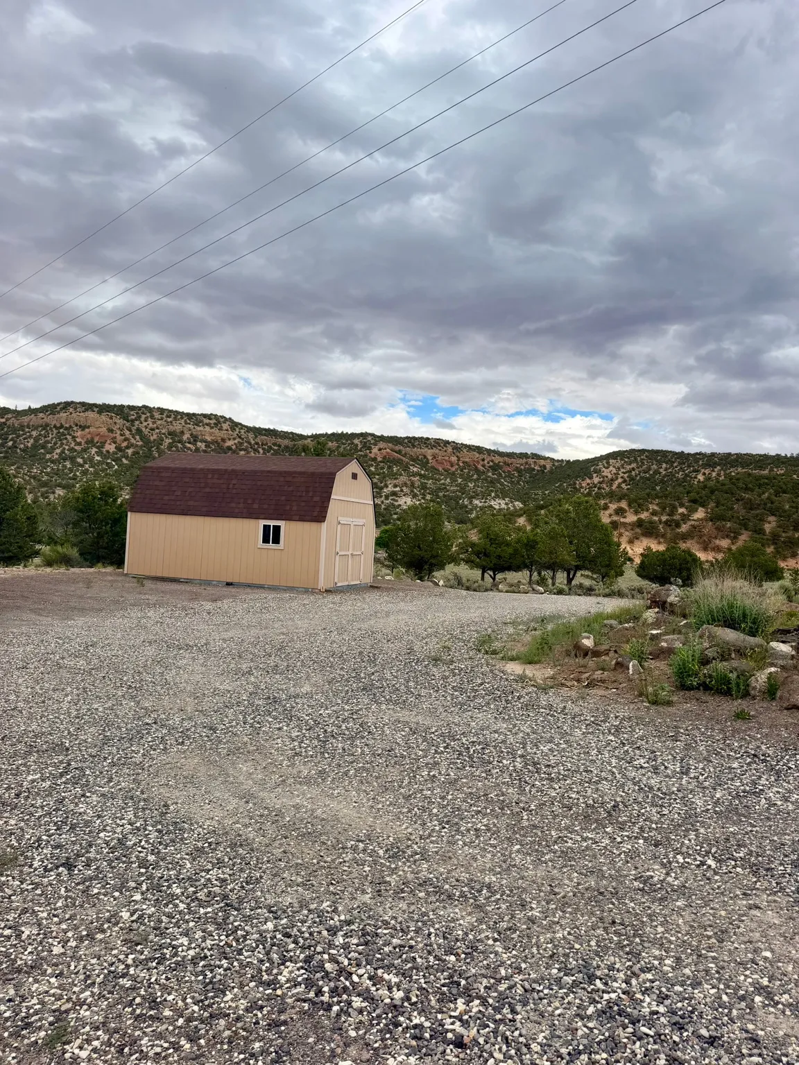 Freestanding storage shed with gambrel roof and a single window, set on a gravel lot