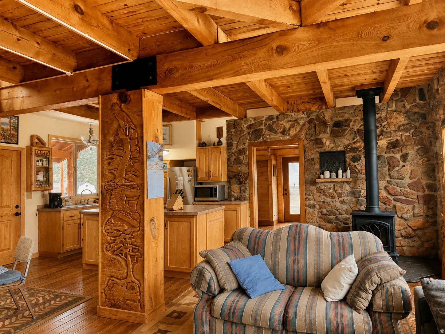 Exposed wood beam ceiling, wood-finish flooring, stone accent wall, black wood-burning stove, and carved wood pillar