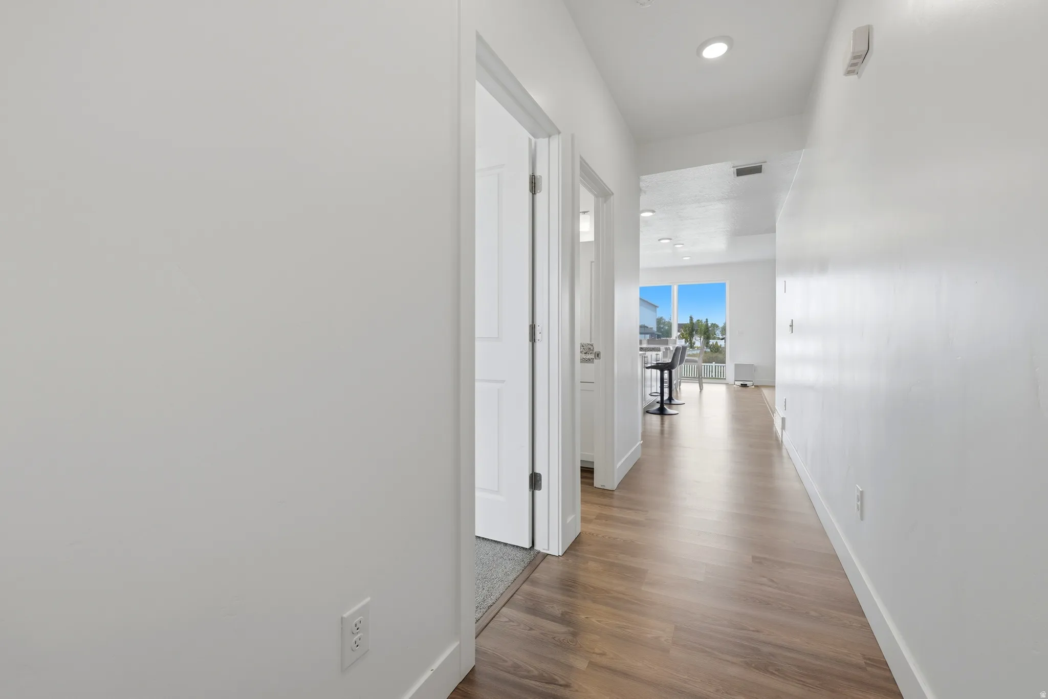 Hallway featuring wood-finish flooring, white walls, recessed lighting, and a sightline to a bright living area with large windows