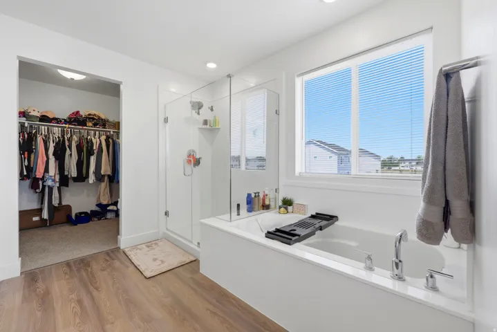 Modern bathroom featuring a built-in soaking tub with chrome fixtures, a glass-enclosed shower, wood-finish flooring, and a spacious walk-in closet with carpeted flooring