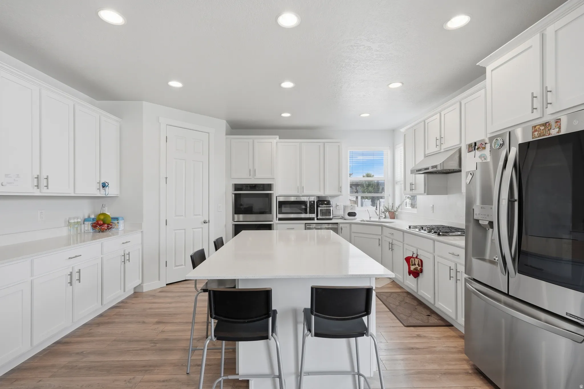 Bright kitchen featuring white shaker cabinetry, stainless steel appliances, a large center island with seating, recessed lighting, and wood-finish flooring