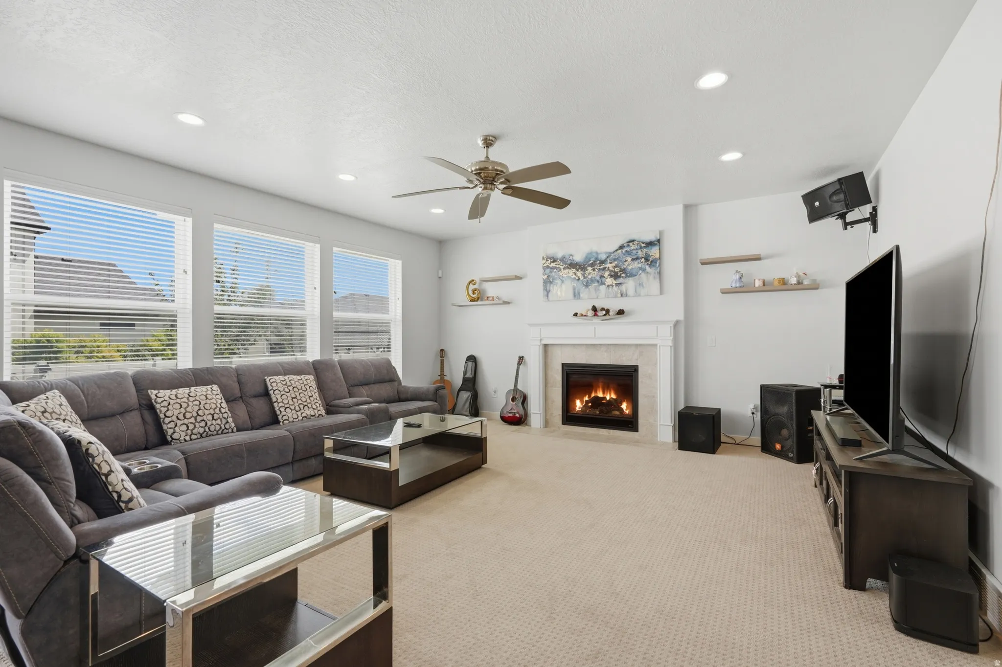 Bright interior space featuring a central fireplace with a white mantel, multiple windows with horizontal blinds, a ceiling fan, recessed lighting, and wall-mounted shelving