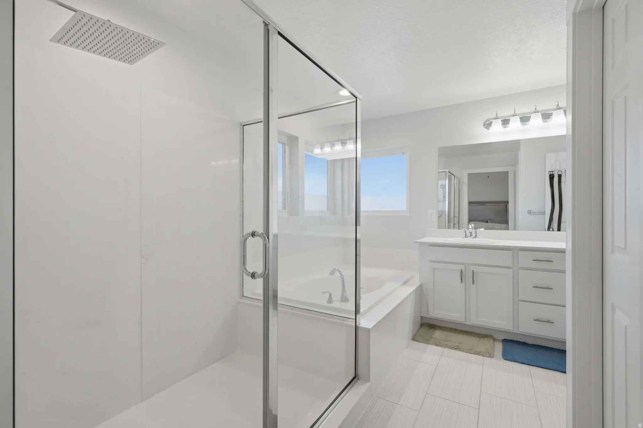 Spacious bathroom featuring a glass-enclosed shower with a rain shower head, a separate soaking tub, a single vanity with a white countertop and base cabinetry, and light-toned tile flooring